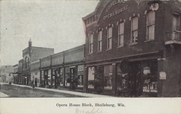 Text on the front reads: "Opera House Block, Shullsburg, Wis." Diagonal view across unpaved street. The building on the right is the red brick, Italianate, two-story Opera House, built in 1882 by the Joseph Copeland family for the city. The ground floor has been home to many businesses. Storefronts and pedestrians are along the sidewalk to the left side of the building.