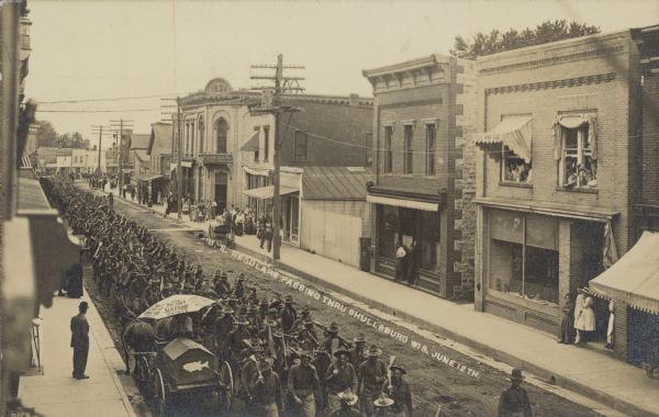 Text on front reads: "U.S. Regulars passing thru Shullsburg Wis. June 12th." Elevated view of a long line of uniformed soldiers, four abreast, marching down an unpaved main street. A crowd is watching from the sidewalk, doorways, windows and horse-drawn vehicles. Many businesses line the street. In the left foreground at the curb is a wagon with a fish logo, and a large umbrella that reads: "Use Patton's Sun-Proof Paints."