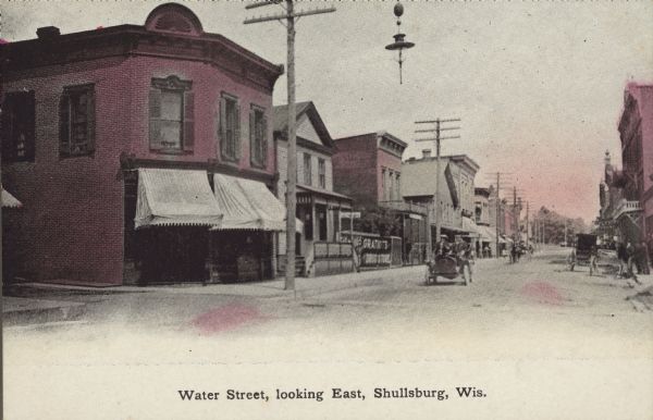 Text on front reads: "Water Street, Looking East, Shullsburg, Wis." People are in an automobile coming up the unpaved street which is lined with businesses. Horse-drawn wagons are hitched at the curb. A sign on the left reads: "Gratiots Drug Store."