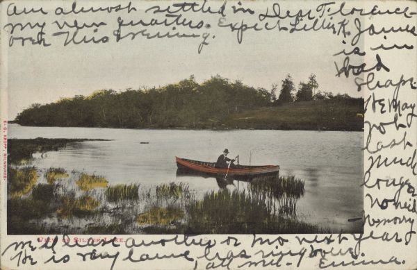 Text on front reads: "View of Silver Lake." A colorized view of a man in a suit rowing a boat on a bay of a lake. Trees and a pasture are on the opposite shore.