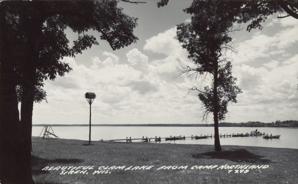 Caption reads: "Beautiful Clam Lake From Camp Northland, Siren, Wis." Two people are in a rowboat and two more are fishing at the end of the pier. Four more rowboats with motors are moored at the pier. Trees, a birdhouse and a swing set are on the lawn at the shore. The far shore is on the horizon.