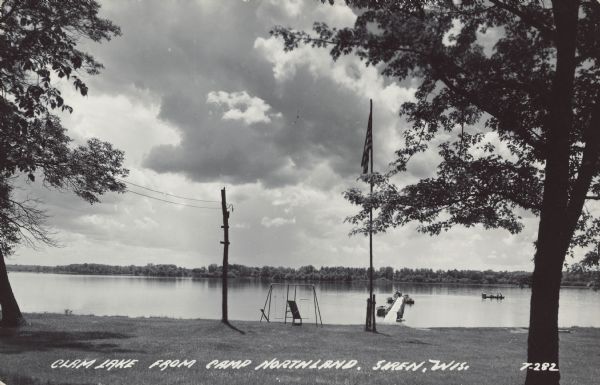 Caption reads: "Clam Lake From Camp Northland, Siren, Wis." Four people are in a rowboat and two more are fishing at the end of the pier. Several more rowboats are moored at the pier, and some have motors. Trees, an American flag and a swing set are on the lawn at the shore. The far shore is on the horizon.