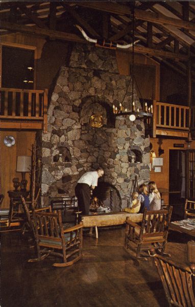 Text on reverse reads: "Hotel du Nord, Sister Bay (Door County) Wisconsin. The giant fireplace, reaching two stories high, is located in the lobby where many pleasant hours are spent. Phone (414)854-4221. AAA." A woman and two girls are sitting on a log bench as a man is tending the fire in a two-story stone fireplace. Above are open trusses, a balcony and mounted bull horns. Many rustic chairs and furniture provide an area for guests.