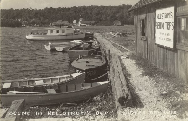 Text on front reads: "Scene at Kellstrom's Dock, Sister Bay, Wis." View of Sister Bay from a boathouse and dock, the sign reads: "Kellstrom's Fishing Trips, 8:00 A.M. to Noon, 1:30 P.M. to 6:00 P.M." Many boats are tied up at the dock. Buildings are on the shoreline across the bay.