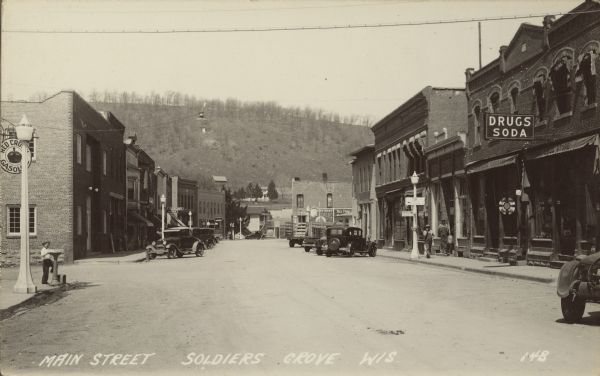 Caption reads: Main Street, Soldiers Grove, Wis." The main street of a small town with businesses on each side. Automobiles and trucks are parked at the curb and pedestrians are on the sidewalks. Some of the signs read: "Red Crown Gasoline," "Drugs, Soda," "Sorensons Furniture," "Columbia Records, "Restaurant" and "Rooms." A tree and grass covered bluff rises beyond the town.
