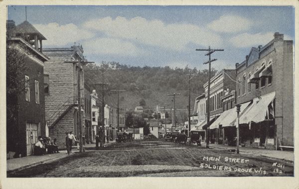 Text on front reads: "Main Street, Soldiers Grove, Wis. 1921." The main unpaved street of a small town with businesses on each side, including a barber, service garage, pool hall and restaurant. Automobiles and trucks are parked at the curb and pedestrians are on the sidewalks. On the left a group of men are gathered on the sidewalk near a drinking fountain which is in front of a brick building with a bell tower. A tree and grass covered bluff rises beyond the town.