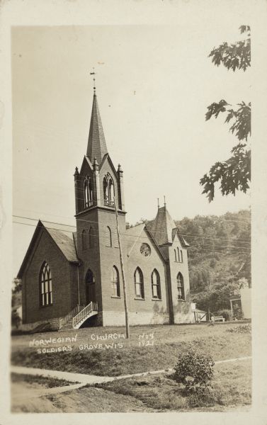 Text on front reads: "Norwegian Church, Soldiers Grove, Wis." View from unpaved road of the front and right sides of the Norwegian Church, which is a brick building with arched stained glass windows, a belfry and a steeple with a weather vane. Below the large arched stained glass window in the front is a bulkhead in the stone foundation. Behind the church on the right is a house. In the foreground is a sidewalk. In the background is a bluff with trees.