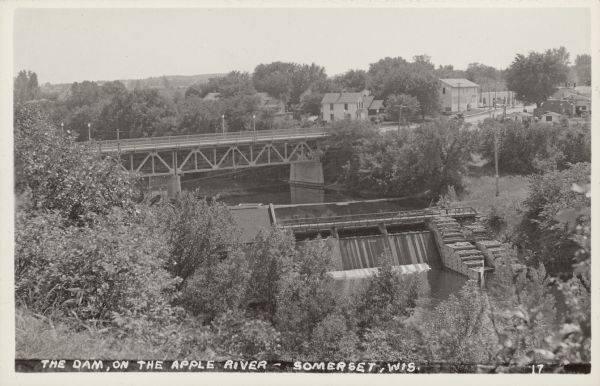 Elevated view of a dam, bridge over the river and part of the town with many trees. Caption reads: "The Dam, on the Apple River - Somerset, Wis."