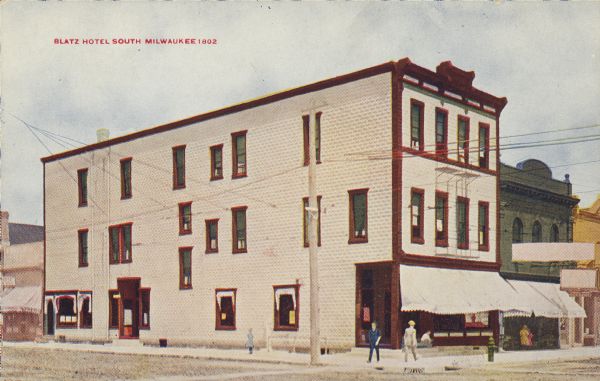Text on front reads: "Blatz Hotel South Milwaukee." Corner view of a three story hotel. The street is unpaved and pedestrians are on the sidewalks. Other businesses are along the sidewalk.