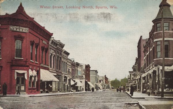 Text on front reads: "Water Street, Looking North, Sparta, Wis." Unpaved street with sidewalks lined with businesses. Signs read, "Monroe County Bank," "Drugs," "Rexall," "Herbst," "Anders" and "Jones Restaurant." There are pedestrians on the sidewalks and horse-drawn vehicles in the street.