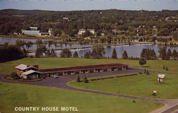 Text on front reads: "Country House Motel." On reverse: "Country House Motel. Northwest's Finest - Highways 53 and 63. Room Phones - Color TV - Air-Conditioned. AAA. Spooner, Wisconsin 54801. Jim & Virginia Woelfel. Phone (715)635-8721." The previous owners were Mr. and Mrs. Lee Hacker. A one-story motel with a two-story building on one end. A large lawn with a few trees and shrubs surround a parking lot. In the background are a series of ponds separated by roads, and beyond are wooded hills and buildings of the town. A water tower is near the top of the hill.