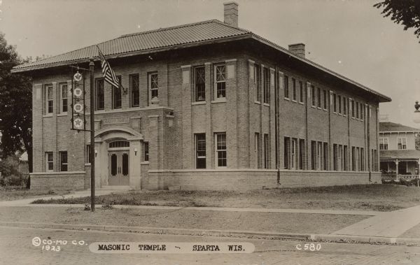 Text on front reads: "Masonic Temple - Sparta, Wis." This Neoclassical building was built in 1923 and faced with tapestry cream brick. In front is a signpost with four masonic symbols. The street is paved with brick.
