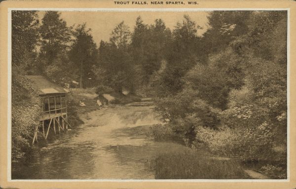 Text on front reads: "Trout Falls, Near Sparta, Wis." Elevated view of river. On the left bank at the base of the falls is an open-sided stilt structure with a partially obscured sign inside that reads: "SHOE". On the bank beyond the structure are two women sitting and viewing the falls. Located 7.7 northeast of Sparta, on the La Crosse River, the falls are surrounded by trees. 