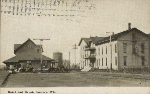 Text on front reads: "Hotel and Depot, Spooner, Wis." A view of the railroad depot and platform on the left, with the hotel on the right. People are on the platform with hand carts carrying goods and baggage. The street in between is made of wood.