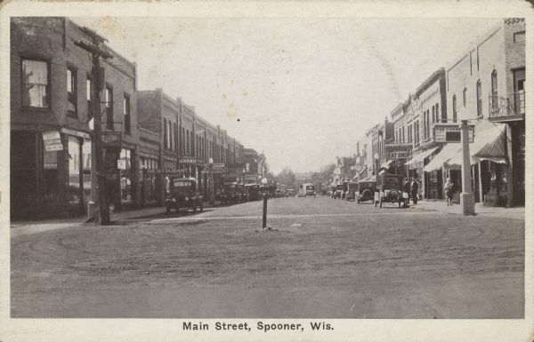Text on front reads: "Main Street, Spooner, Wis." Unpaved main street with many businesses on both sides. Automobiles and trucks are on the street or parked at the curb and pedestrians are on the sidewalks. Some of the signs read: "Restaurant", "Bakery", Grand" and "Benson's Eat Shop". 