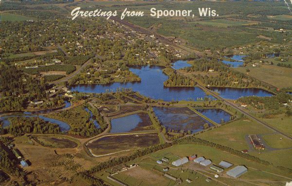 Text on front reads: "Greetings from Spooner, Wis." Text on reverse: "Looking north on this aerial view one can see the State Fish Hatchery in the foreground of the picture, which is just south of the city limits. Musky and other species are reared here by the state for stocking the many lakes in the area." Aerial view of the town and the Yellow River Flowage.