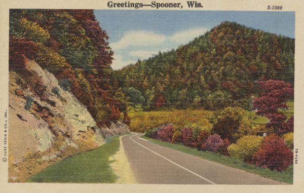 Text on front reads: "Greetings - Spooner, Wis." A country road descends into a valley between tree-covered hills. On the right are exposed rock faces at the foot of the bluffs. The trees are showing autumn color.
