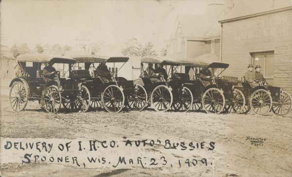 Text on front reads: "Delivery of I.H. Co. 'Auto' Buggies. Spooner, Wis. Mar. 23, 1909." Men are posing while sitting in the driver's seats of five International Harvester Auto Buggies on a dirt road in front of the Spooner Lumber Company. Trees and dwellings are in the background.