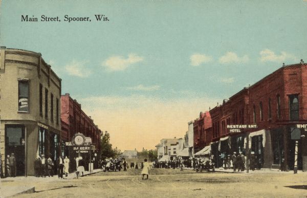 Text on front reads: "Main Street, Spooner, Wis." Unpaved main street with many businesses on both sides. Automobiles and trucks are on the street or parked at the curb and pedestrians are on the sidewalks. One woman is crossing the street. Some of the signs read: "Restaurant", "Hotel", "Bakery" and "Lunches". 