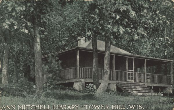 Text on front reads: "Ann Mitchell Library-Tower Hill, Wis." A one-story, wooden building with a wrap-around veranda and broad front steps surrounded by trees. This property became part of Tower Hill State Park in 1922. The library has ties to Lutie Eugenia Stearns, Wisconsin library pioneer, and Jenkin Lloyd Jones, a prominent Unitarian Minister.