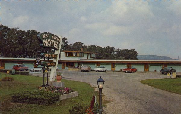 Text on reverse reads: "Rest Haven Motel. Air Conditioned - Free TV - Room Phones. Free Coffee - Free Golf. 'The Best In Rest' John and Catherine Michels. Phone 588-4191. Intersection Hwy 14 and State 23. Spring Green, Wisconsin." A roadside motel with cars parked in front of each unit, with lawn chairs on the right and left. A couple is walking into the office. The sign for the motel is in the foreground with landscaping and reads: "Rest Haven Motel. Family Units. No Vancancy. AAA". Trees and bluffs are in the background. The motel was designed by Taliesin-affiliated architect, J.C. Caraway and constructed by Kramer Brothers Builders in 1952. It was renamed "The Usonian Inn" in 1991.