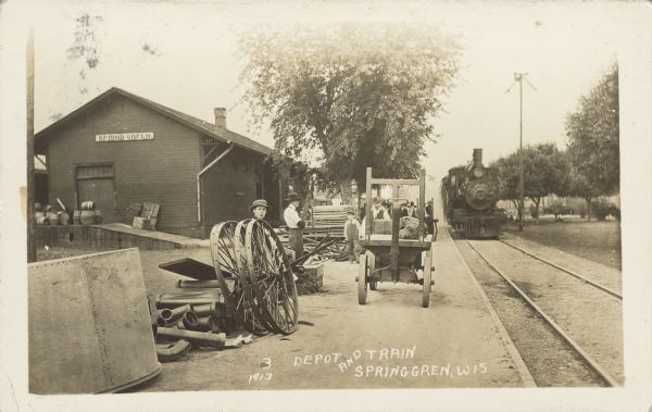 Text on front reads: "Depot and Train, Spring Gren [sic], Wis." A train station with the depot on the left and a locomotive on the right. Adults and children are standing in the central area, with cargo and baggage on the ground, as well as carts. Some of the cargo includes wagon wheels, barrels and pipes.