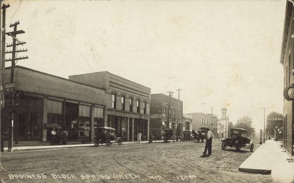 Text on front reads: "Business Block, Spring Green, Wis." An unpaved street with automobiles and horse drawn vehicles, many parked at the curb. Pedestrians are on the sidewalks and street in front of many businesses. Storefront signs read: "Garage, Free Air", "Sherwood Bros. Hardware and Furniture" and "Restaurant".