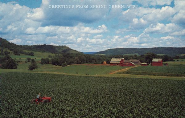 Text on front reads: "Greetings from Spring Green, Wis." On the reverse it reads: "Lush Farm Land." Elevated view of a farmer on a tractor in a large corn field. In the background are farm buildings, a haystack and pastures. In the far distance are tree-covered bluffs.