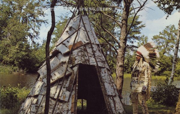 Text on front reads: "Greetings from Spring Green." On the reverse it reads: "Be It Ever So Humble..." A Native American man dressed in indigenous clothing is posing with a birch bark tipi. In the background is a river and trees.