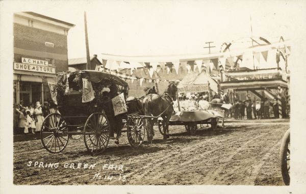Text on front reads: "Spring Green Fair." A parade through a town on an unpaved road. A horse-drawn buggy is decorated with "Erin Go Bragh" flags and foliage. The horse-drawn wagon in front of it has people sitting on it. The buildings are decorated and above the streets are  banners strung on wires. Many spectators are watching from the sidewalks. On the left is the "A.C. Hare Shoe Store."