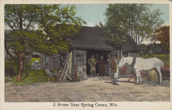 Text on front reads: "A Scene Near Spring Green, Wis." Two men are standing in the open doorwway, and one man is standing with a white horse in the yard of a building, with equipment and wood stacked alongside the left side of the building. Another building is in the background on the left, and a field is on the right.