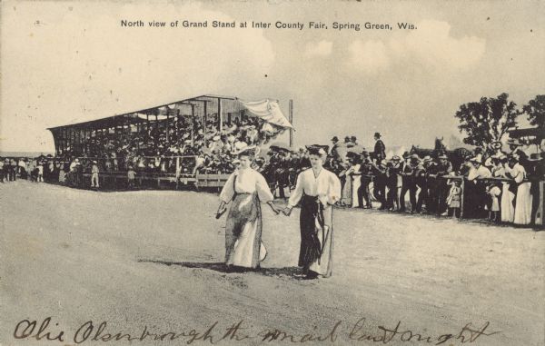 Text on front reads: "North view of Grand Stand at Inter County Fair, Spring Green, Wis." Two women are walking on the track in the foreground. In the background is a crowd on and around the grandstand at a fair. Horse-drawn vehicles are behind the crowd.