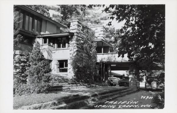 Text on front reads: "Taliesin, Spring Green, Wis." Stone walls, steps and windows are landscaped with trees and shrubs, on the northeast elevation of the studio and balcony off the studio of Taliesin, home of Frank Lloyd Wright. Automobiles are parked under the balcony.