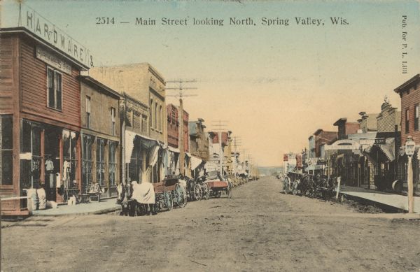 Text on front reads: "Main Street Looking North, Spring Valley, Wis." View down center of unpaved street with sidewalks, lined with businesses. Horse-drawn vehicles are parked at the curb and pedestrians are on the sidewalks. Some of the signs read: "Hardware & Stoves," "Drug Store," "K. Peterson Boots & Shoes" and "K. Hanson" and "Bakery."