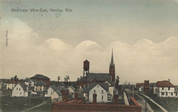 Caption reads: "Bird's-eye View-East, Stanley, Wis." Elevated view from a rooftop, with the flat roof of a brick building in the foreground. In the center background are a church and a water tower surrounded by houses. The streets are unpaved and lined with sidewalks.