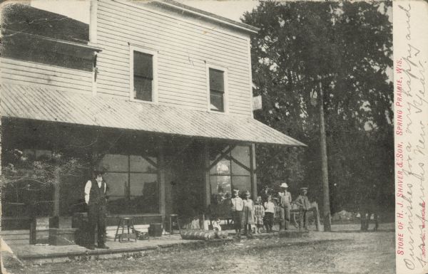 Text on front reads: "Store of H.J. Shaver & Son, Spring Prairie, Wis." A clapboard store with large front windows. A group is posing in front, with a man on the left, and a group of children with a dog on the right. Merchandise is on display behind them. A wagon and trees are on the right.