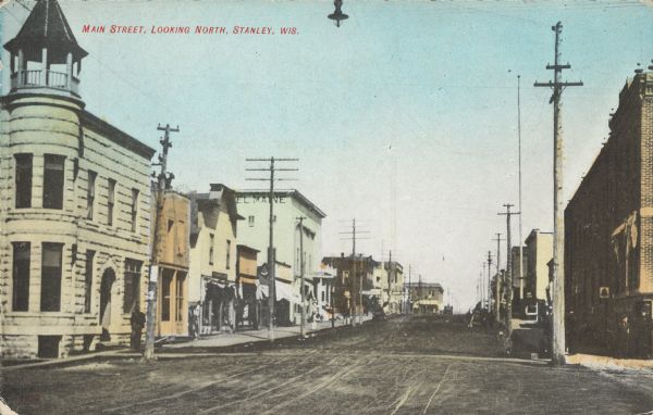 Caption reads: "Main Street, Looking North, Stanley, Wis." View up unpaved street lined with businesses. Pedestrians are on the sidewalks. The Hotel Maine is five or six buildings down on the left.