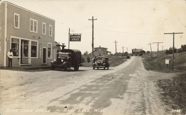 Text on front reads: "Star Lake Store - Star Lake, Wis." J. Mykleby & Son General Merchandise store on an unpaved road with two other dwellings in the distance. The Star Lake Post Office and a Standard Service Station are also located in the same building. A military style truck and an automobile are parked in front. One man is standing on the cement slab smoking a cigarette and another man is talking to the driver of the automobile.