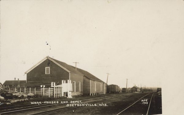 Text on front reads: "Ware-houses and Depot, Stetsonville, Wis." View across railroad tracks towards a train station with a depot, warehouses, cattle loading pen and a pile of logs.