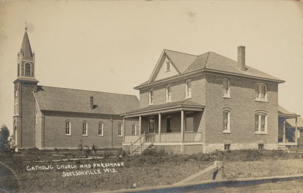 Text on front reads: "Catholic Church and Parsonage, Stetsonville, Wis." Both buildings are built of brick with arched windows, and a log cabin is behind the parsonage on the right.