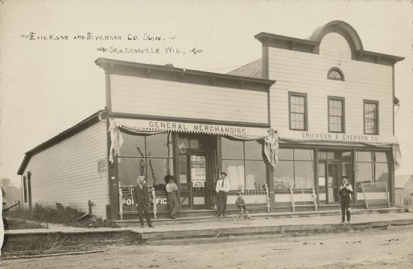 Text on front reads: "Erickson and Evenson Co. Store. Stetsonville, Wis." Four men and a boy on a tricycle pose on the boardwalk in front of a General Store. A woman is standing just inside the door on the right. Several metal bed frames are on display, leaning against the  show windows in front. The awnings are drawn back with the signs on the front reading: "General Merchants" and "Erickson & Evenson Co." A poster in the window announces that the "Minneapolis Keystones", a black baseball team, will be playing on September 13-14.