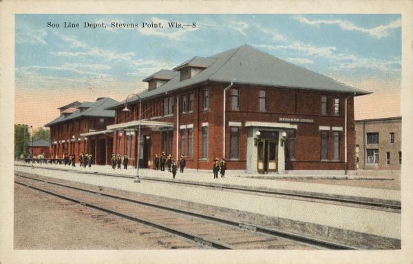 Text on front reads: "Soo Line Depot, Stevens Point, Wis." The depot was built in 1918 of brick in the Prairie School style. There are pedestrians on the platform and tracks in the foreground. Another building is in the background on the right.