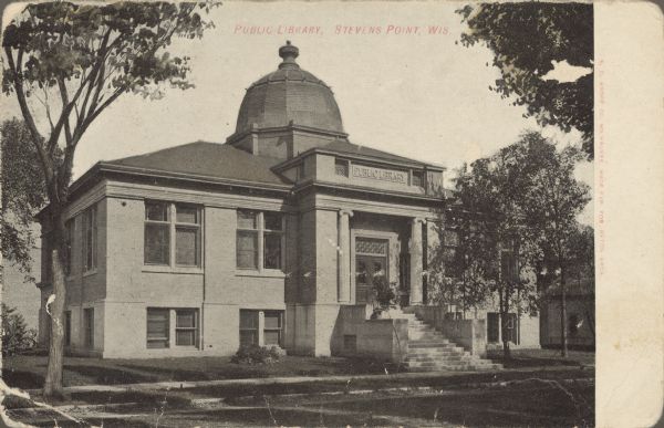 Text on front reads: "Public Library, Stevens Point, Wis." Exterior of the Carnegie Public Library, built in 1903 after the receipt of a 1902 Carnegie Grant. A set of stairs leads from the sidewalk to the main entrance with two Ionic columns on either side of the double doors. The library building is topped by a segmented dome. Trees and a lawn surround the building.