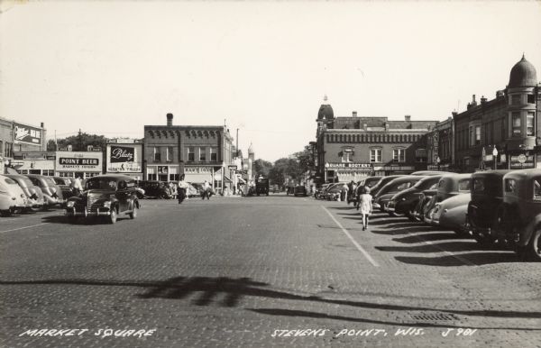 Text on front reads: "Market Square, Stevens Point, Wis." A large, brick paved area, with parking for automobiles, surrounded by businesses. Pedestrians are walking on the square. Some of the signs on the storefronts read: "Miller High Life Beer", "Harness Leather Goods", "Square Deal Tavern", "Drink Point Beer, Barney's Tavern", "Blatz, Wanta's Tavern", "Bartig's", "Square Bootery, Smart Shoes" and "Domack Clothing Store".