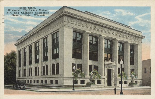 Text on front reads: "Home Office, Wisconsin Hardware Mutual, Fire and Casualty Companies, Stevens Point, Wis." Exterior view from intersection of the Hardware Mutual Insurance office building. The Wisconsin Retail Hardware Association and the Fire and Casualty Company opened the new three-story building on August 2, 1922. The front of the building has stone planters with flowers in the ground level windows and stone planters with lampposts at the sidewalk. The entrance is very ornate with stone relief decorations. An automobile is parked on the left.