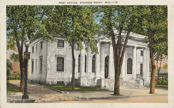 Text on front reads: "Post Office, Stevens Point, Wis." Exterior of the stone building constructed in the classical revival style, built in 1919. Stairs lead to the front doors beyond a row of Doric columns. The street and driveway are paved with brick. Mature trees are in the foreground.