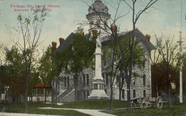 Text on front reads: "Portage Co. Courthouse, Stevens Point, Wis." Exterior of the Portage County Courthouse, built in 1868. Stairs lead to the main entrance of the stone building, and a cannon stands on the lawn on the right. A pavilion is on the left and trees surround the building. In the center is the Portage County G.A.R. Memorial. This monument was dedicated on September 2, 1890 to honor the Civil War Union veterans from Portage County. The granite memorial features a sculpture of a Union soldier holding a flag and grasping his sword at the top of a tall obelisk.