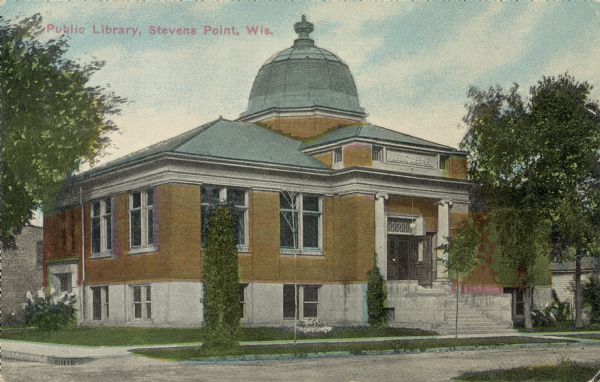 Text on front reads: "Public Library, Stevens Point, Wis." Exterior of the Carnegie Public Library, built in 1903 after the receipt of a 1902 Carnegie Grant. A set of stairs leads from the sidewalk to the double doors of the main entrance flanked by two Ionic columns. The library building is topped by a segmented dome. Trees, shrubs and flowers surround the building.