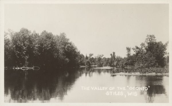 Text on front reads: "The Valley of the 'Oconto', Stiles, Wis." The shoreline is lined with trees. The Oconto River flows into Green Bay.