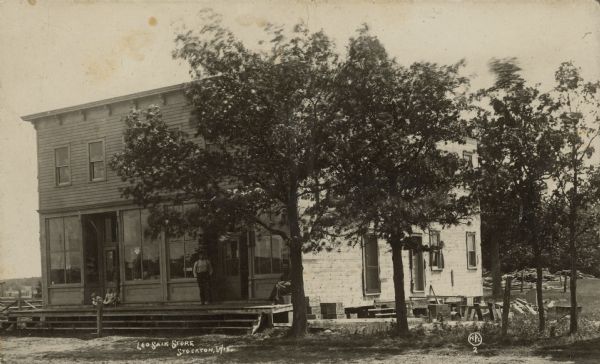 Text on front reads: "Leo Saik Store, Stockton, Wis." Two men and three children pose on the elevated boardwalk in front of a two-story building with a flat roof. The front of the store has large show windows, and two entrances. There are entrances to the building on the right side. Large trees and woodpiles are on the right.